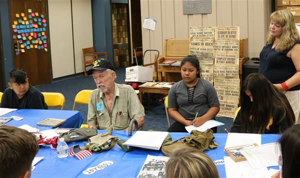Junior high students sit around a table, listening to World War II veteran speak. A woman stands nearby. Junior high students sit around a table, listening to World War II veteran speak. A woman stands nearby.